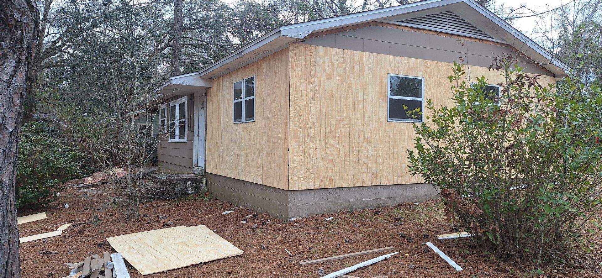 Small house under construction with exposed plywood siding in a wooded area, with building materials in the foreground