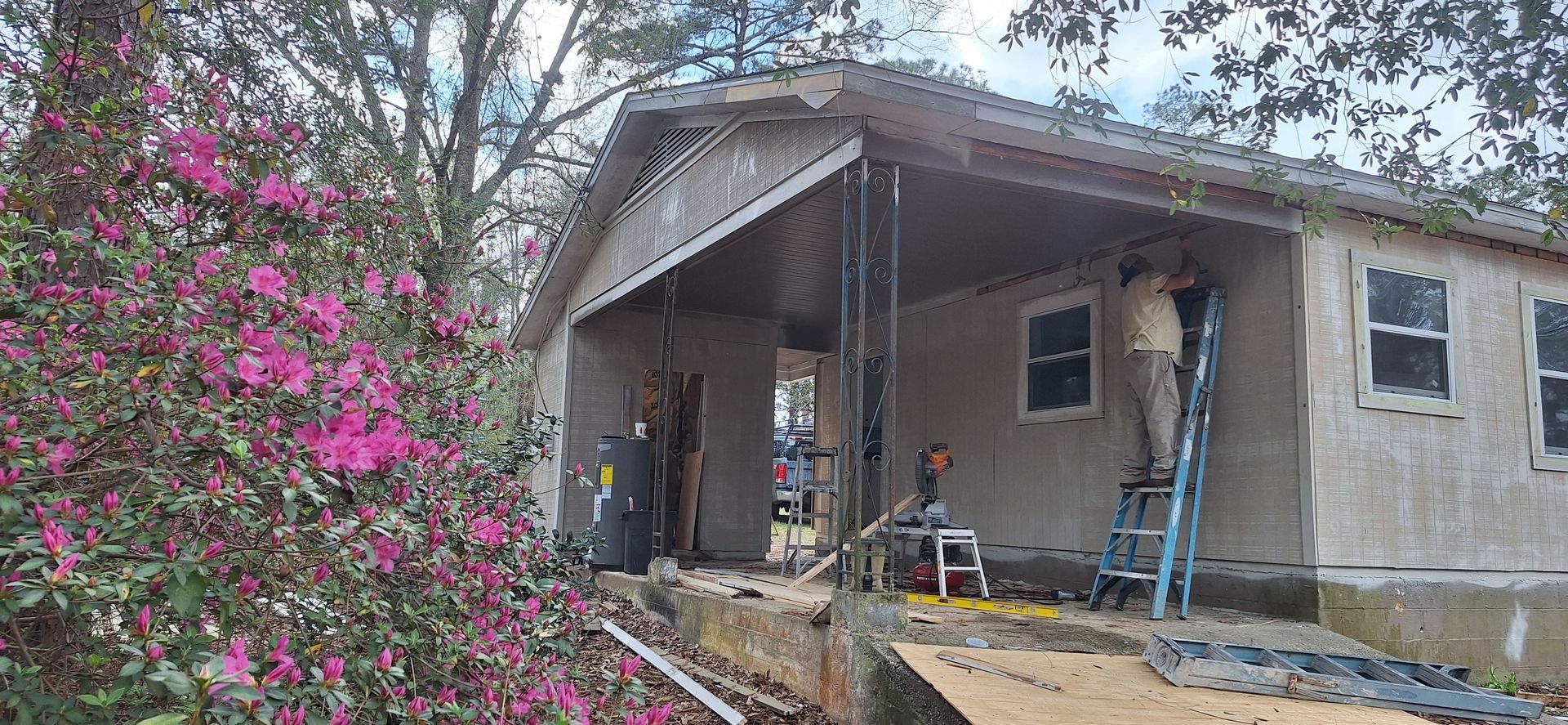 House under renovation with workers on a porch beside blooming pink flowers