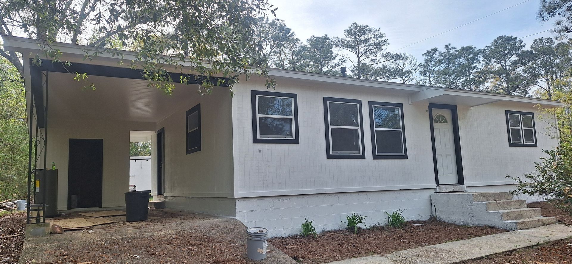 White single-story house with dark trim, front porch, and unfinished exterior in a wooded yard