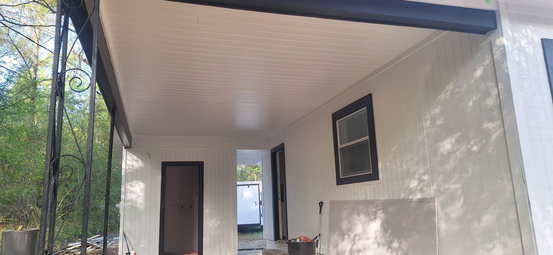 Small porch entry with beige walls, a dark door, and a window, surrounded by trees and greenery.