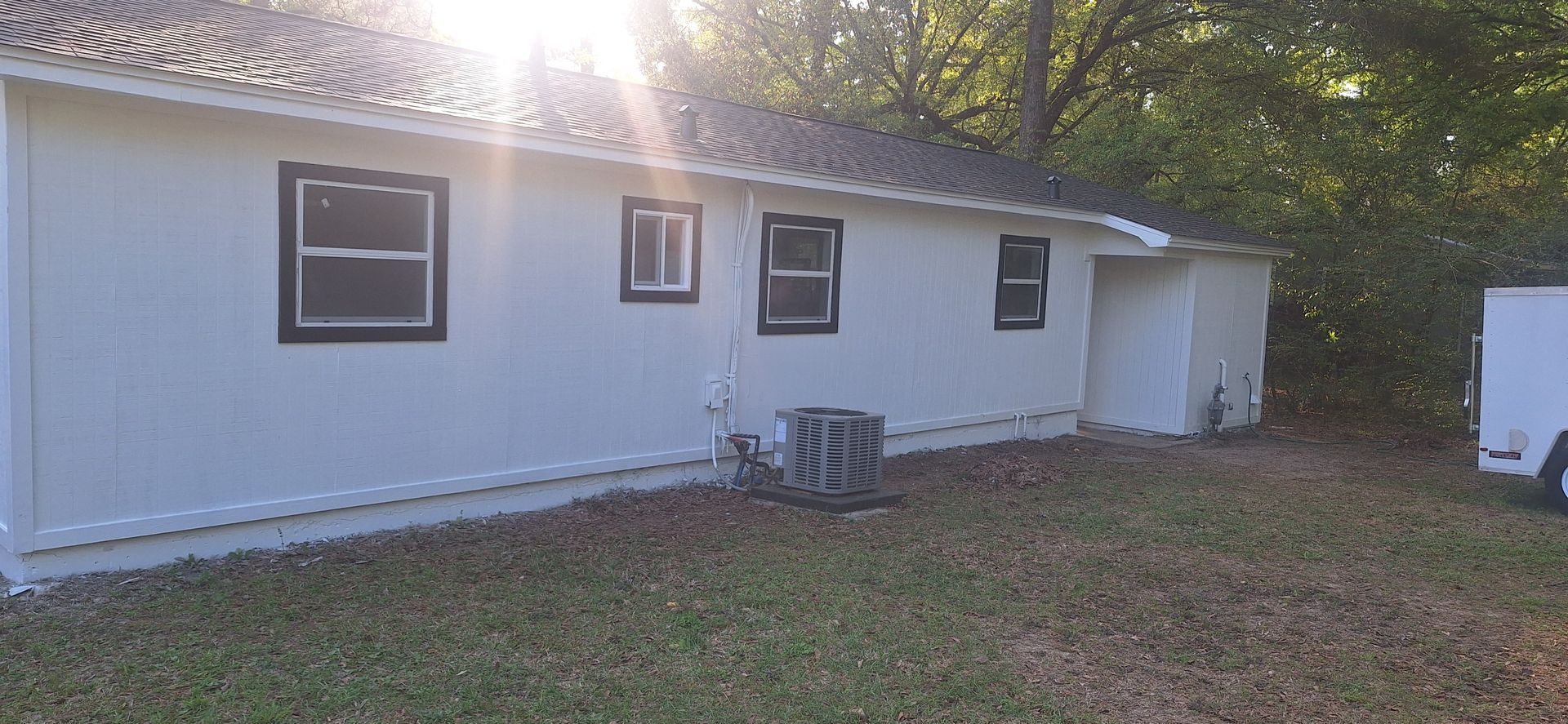 White house exterior with three windows, a small utility unit, and a sunlit yard with trees behind it