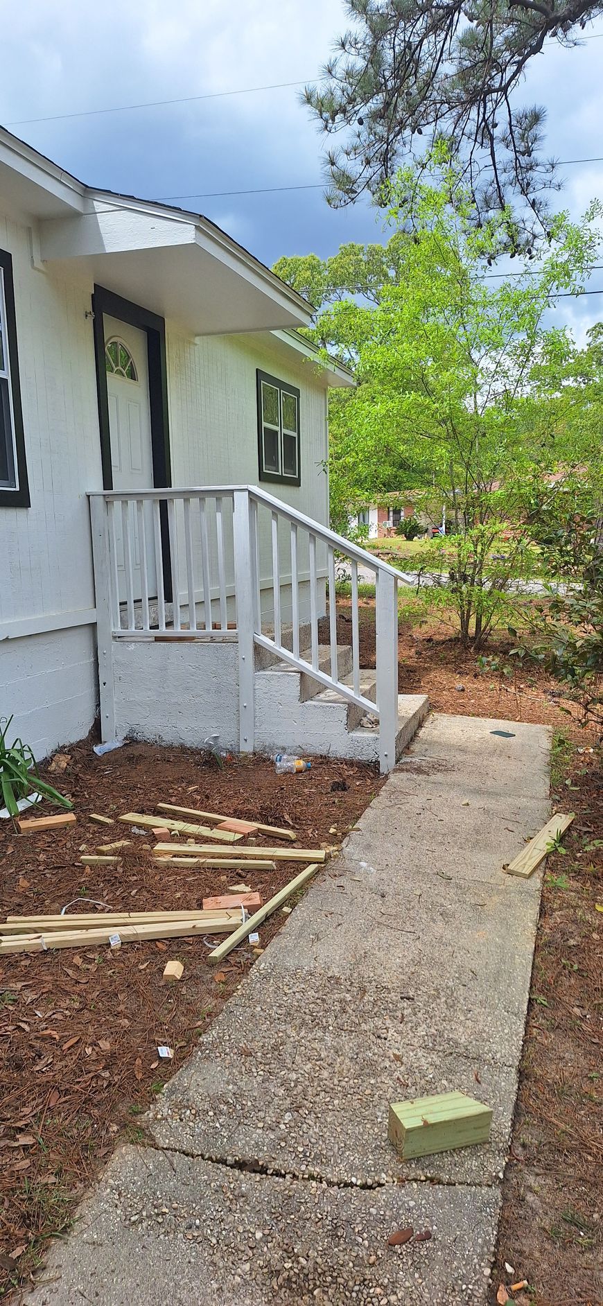 Concrete walkway beside a white house, leading past a small porch with white railing and green trees