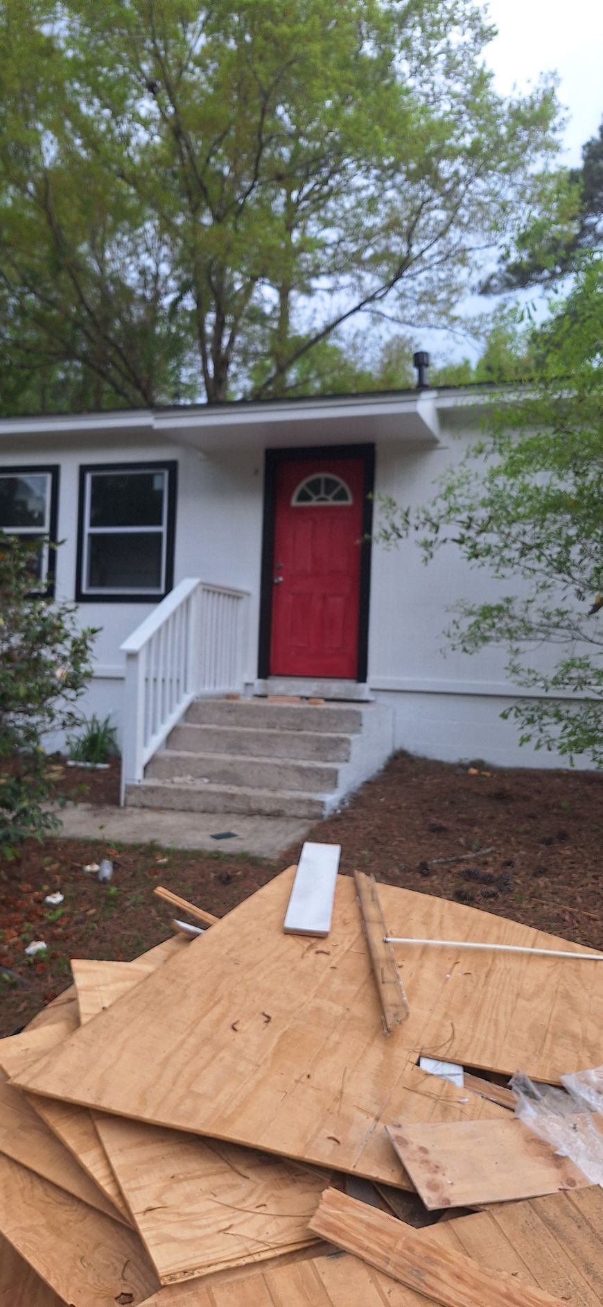 Front of a white house with a red door, white steps, and plywood piled in the yard.