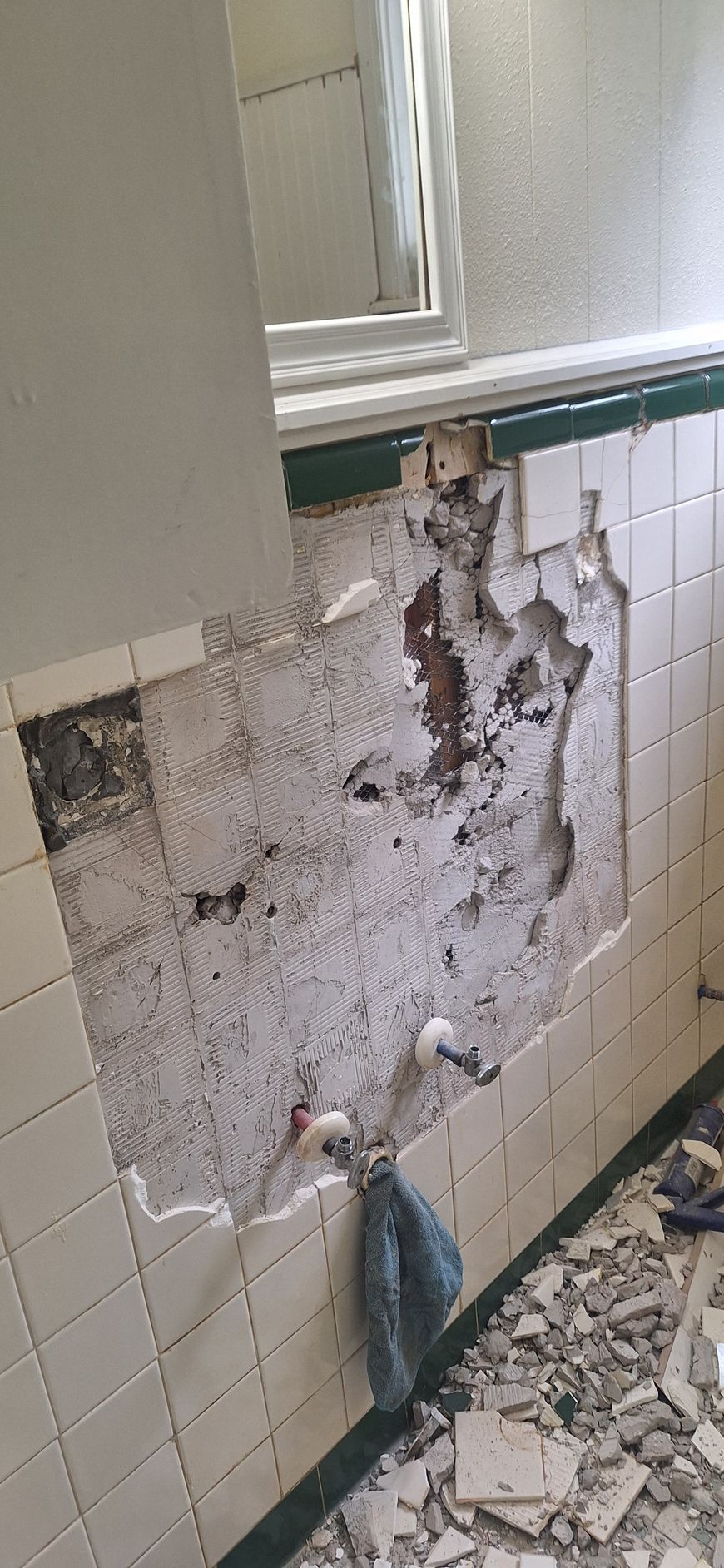 Damaged bathroom wall with broken tiles, exposed plaster, and debris on the floor
