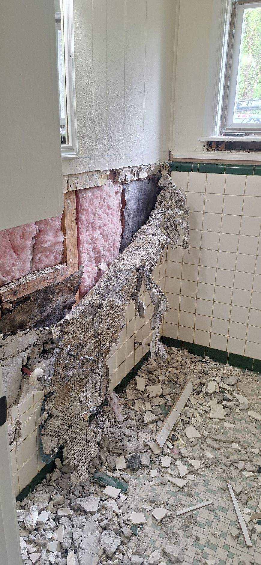Demolished bathroom wall with exposed insulation, torn tile, and debris on the floor near a window