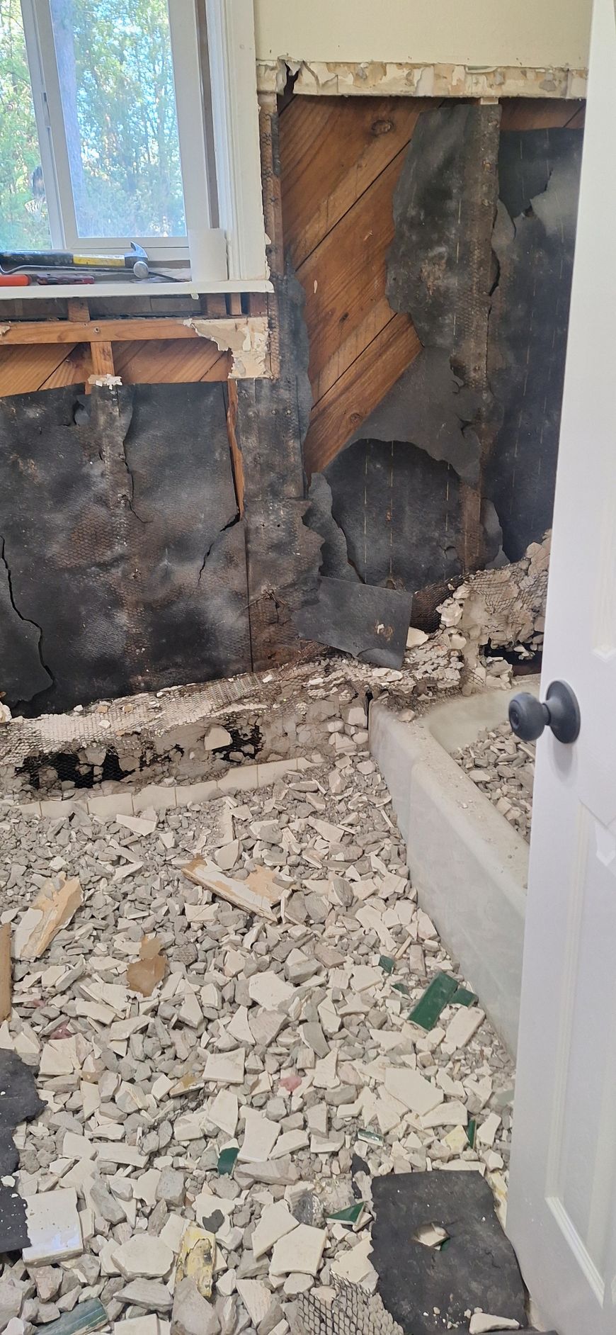 Damaged room wall with exposed insulation, debris-covered floor, and open white door near a window