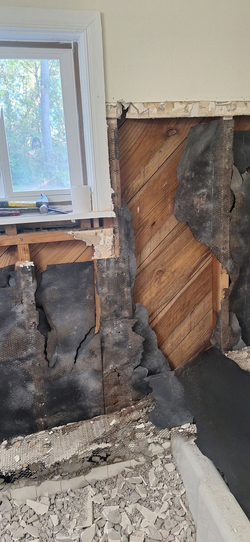 Damaged interior wall with exposed wood framing and insulation beside a window and rubble on the floor