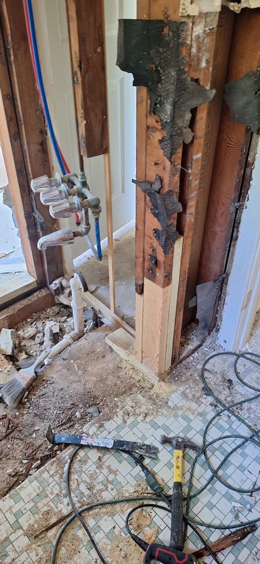 Partially demolished wall exposing plumbing pipes, with debris and tools on the floor