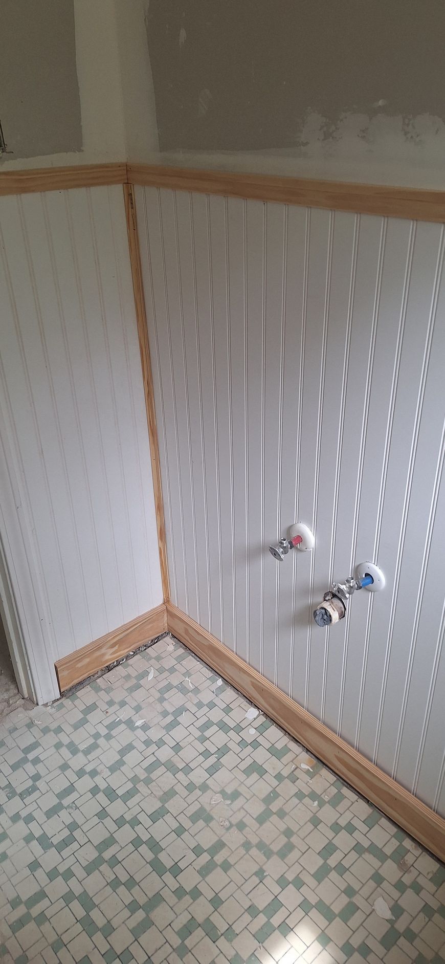Empty tiled bathroom corner with wood trim and exposed plumbing fixtures on the wall