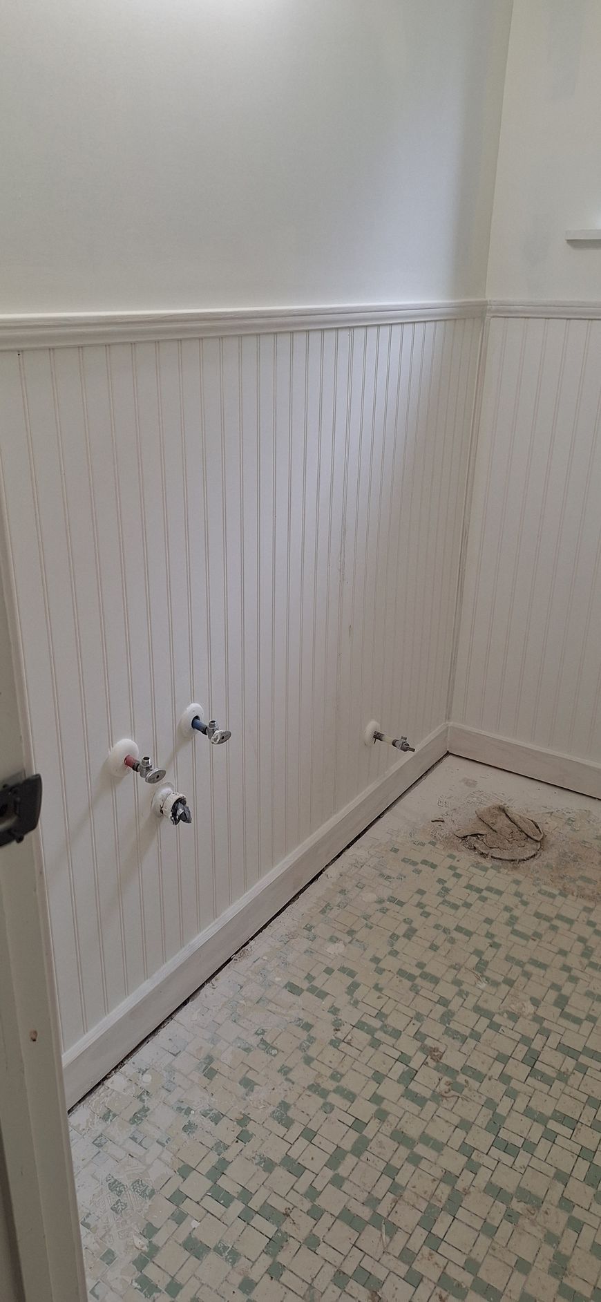 Empty bathroom corner with white beadboard walls, exposed pipes, and a patterned floor.