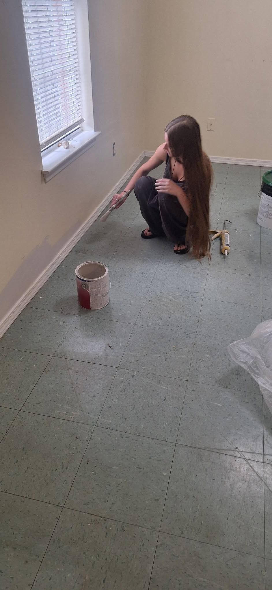 Person kneeling on a gray floor near a window, painting a baseboard with a brush and paint bucket nearby