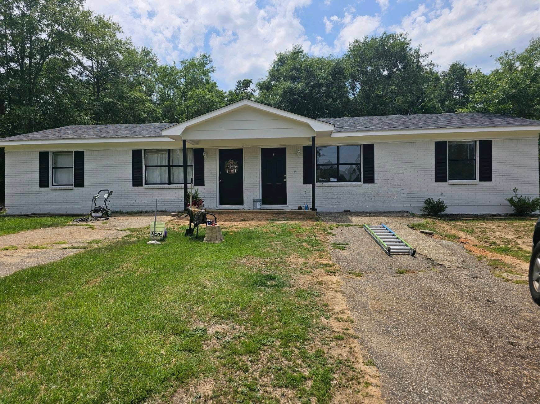 Single-story white house with black shutters, front porch, and a gravel driveway under a cloudy sky