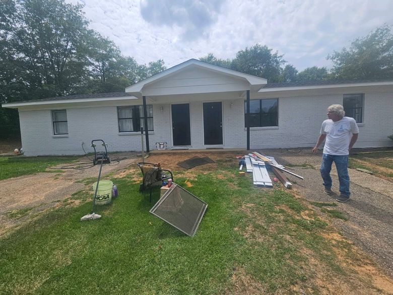 Damaged house exterior with debris in the yard and a person standing nearby