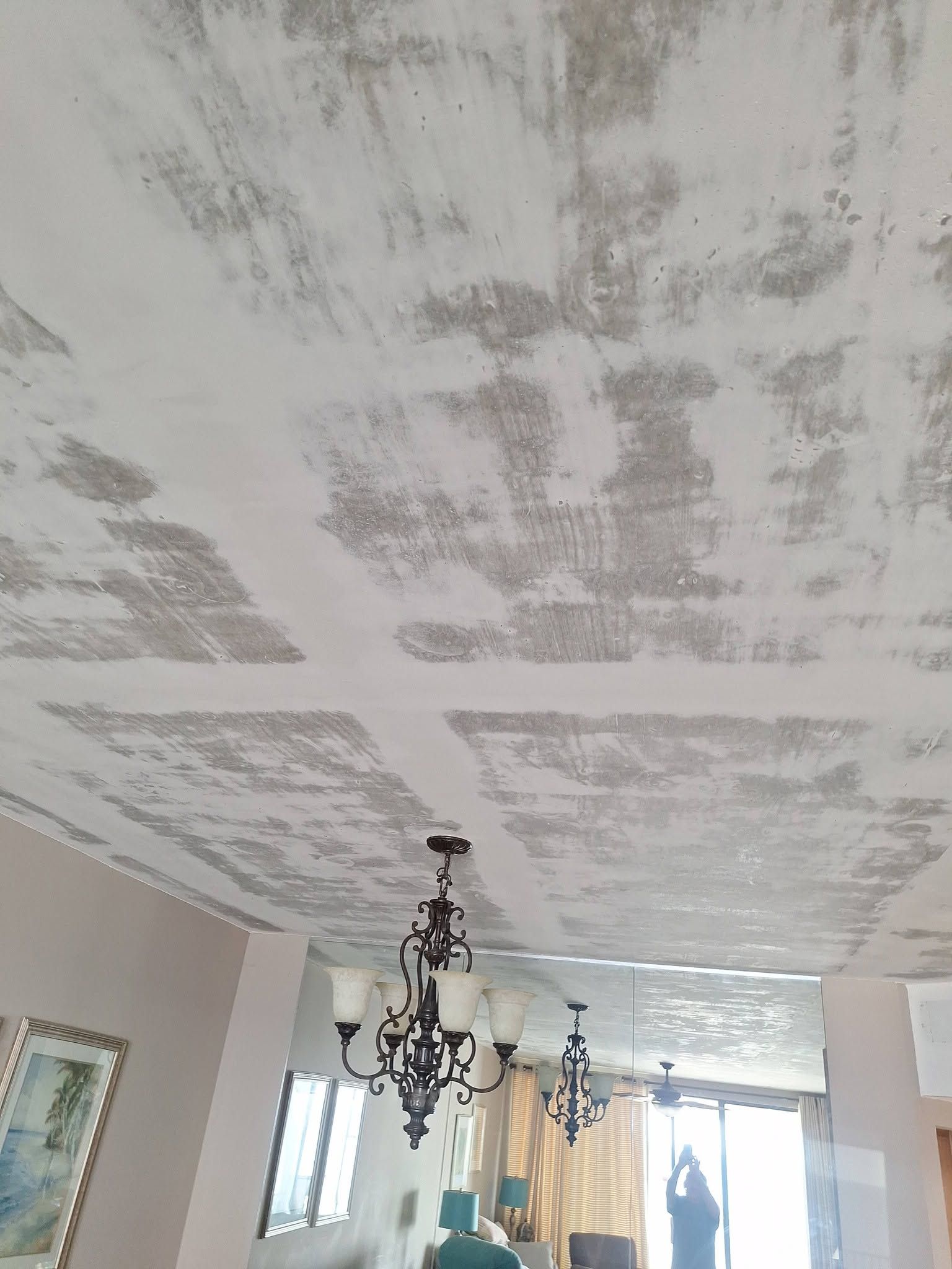 Living room ceiling with patchy plaster repair above a chandelier and windows