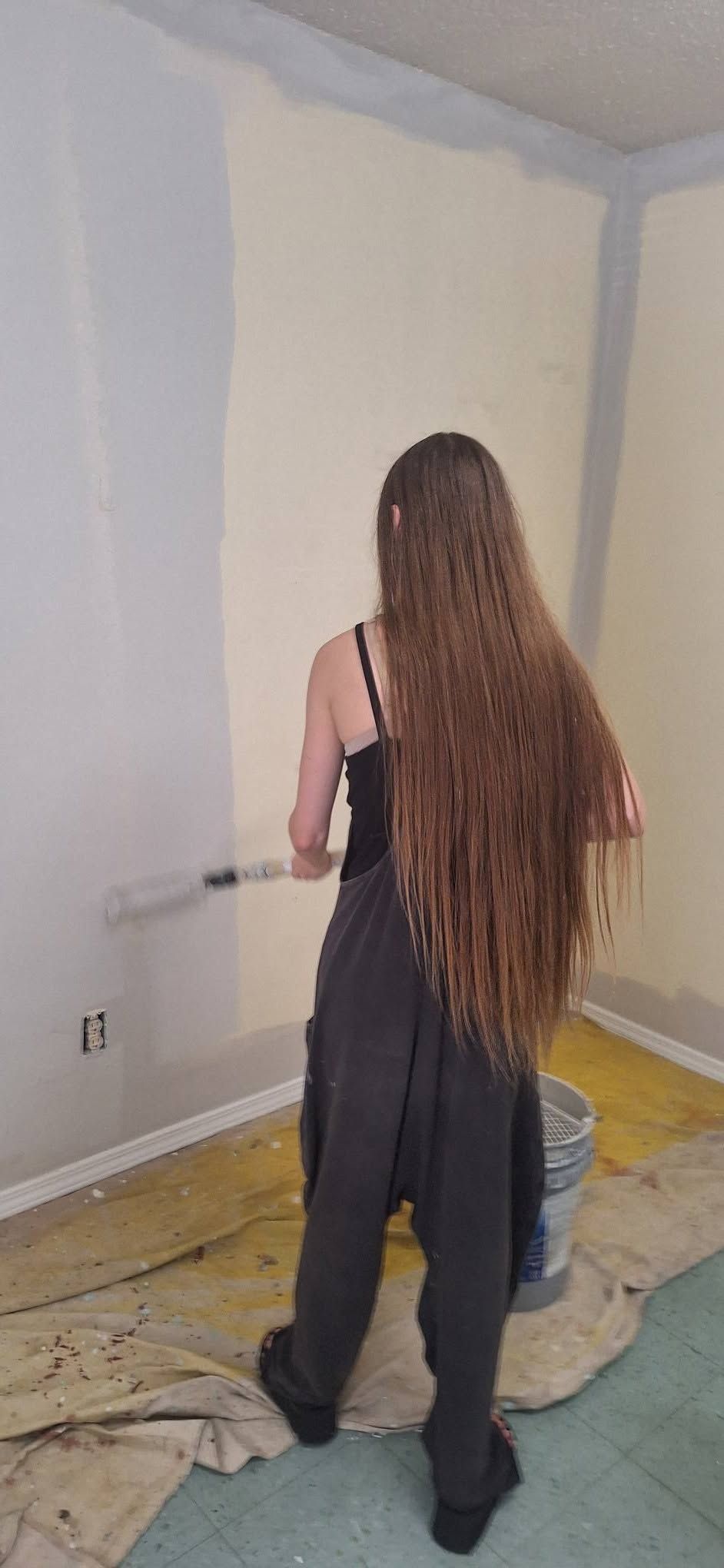 Person standing in a mostly empty room, painting or patching the wall near the floor.