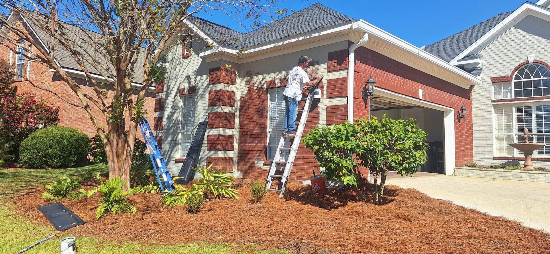 Worker on a ladder painting a brick house exterior beside a garage in a landscaped yard