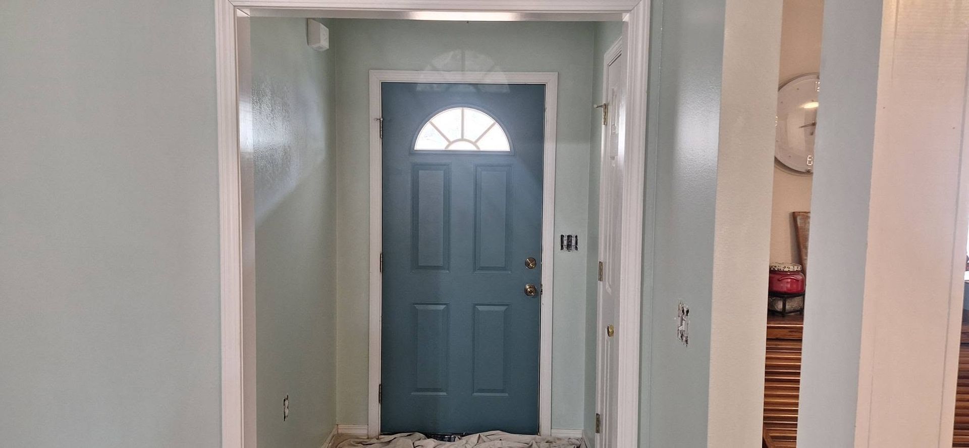 Hallway facing a blue front door with a half-moon window, framed by white walls and doorways.