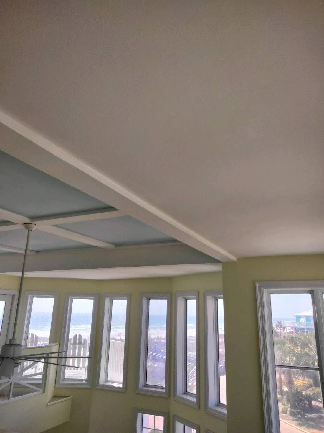 Sunlit room with large windows, pale green walls, and a ceiling fan overlooking trees and buildings
