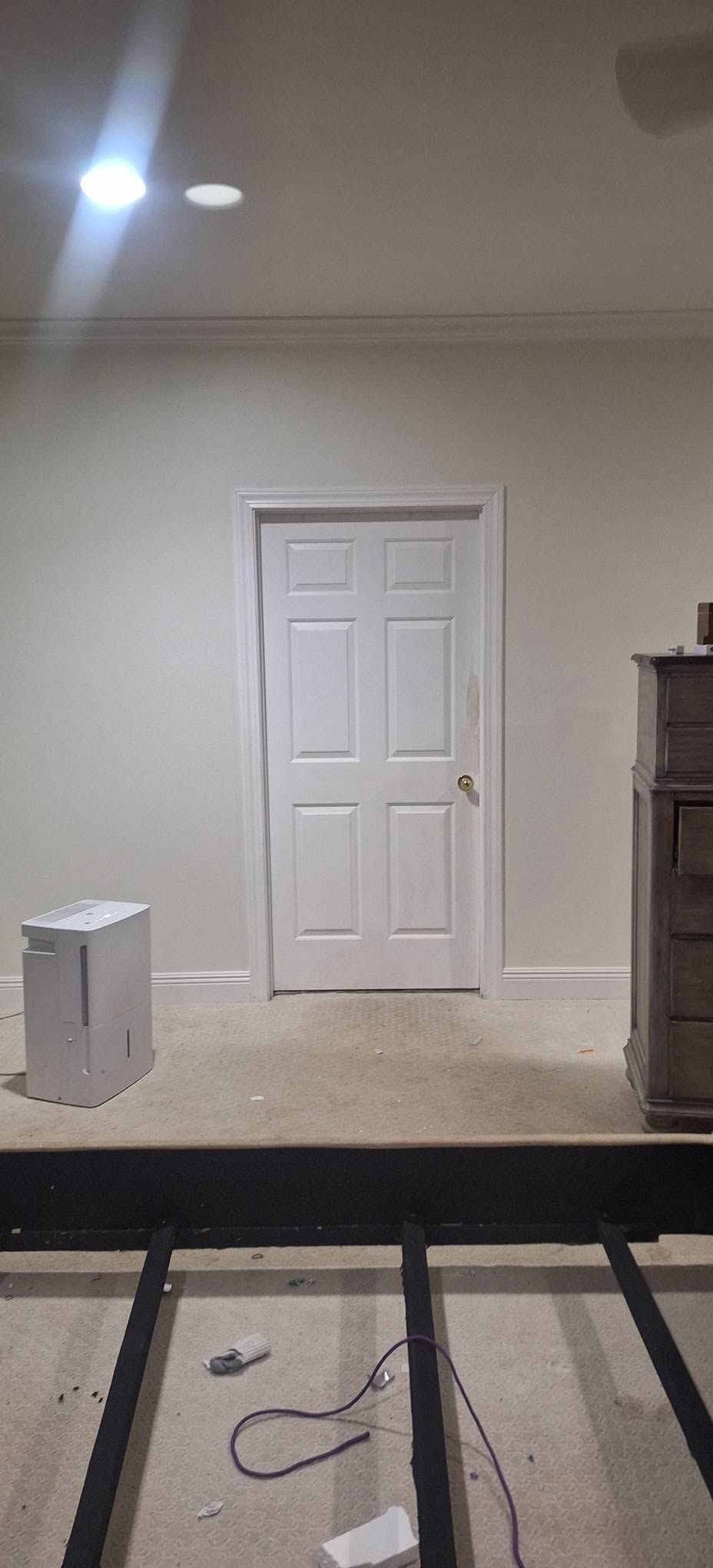 Empty room with white door, beige walls, ceiling light, and exposed floor framing in foreground