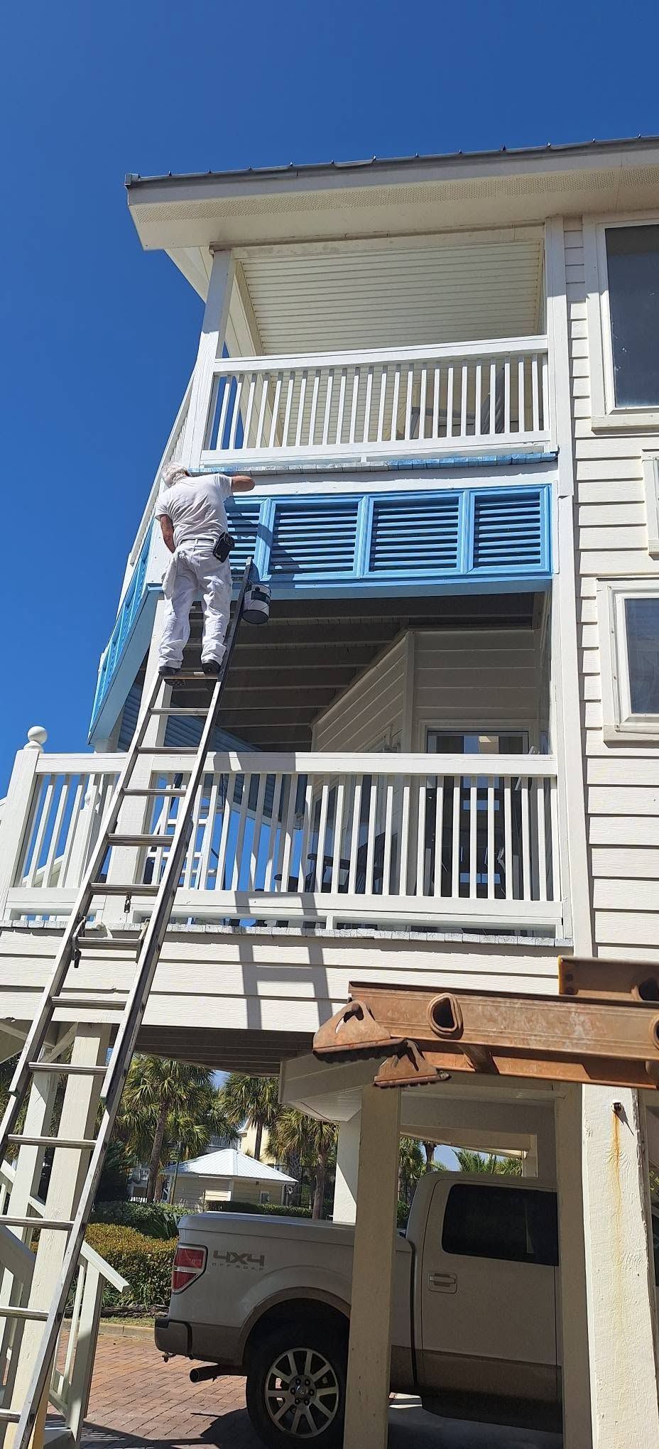 Person on a ladder painting a blue shutter on a white house porch