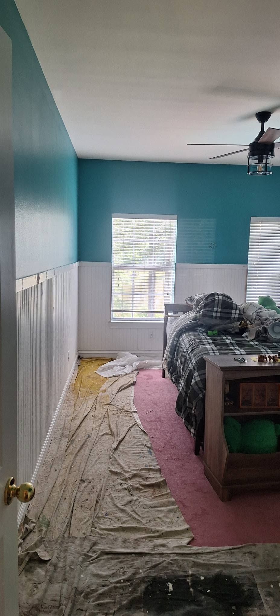 Small bedroom with teal walls, white wainscoting, two windows, and a bed covered with blankets and bedding supplies.
