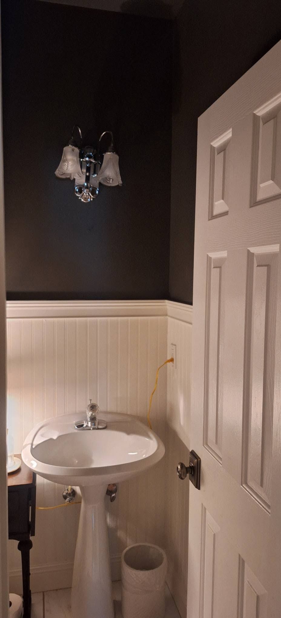 Small bathroom with pedestal sink, dark walls, white wainscoting, and an open door.