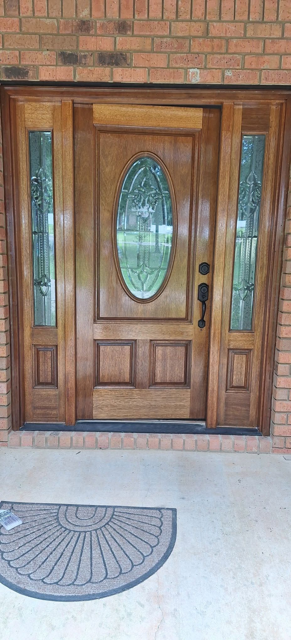 Wooden front door with oval glass panel and side windows beneath a brick wall and doormat.