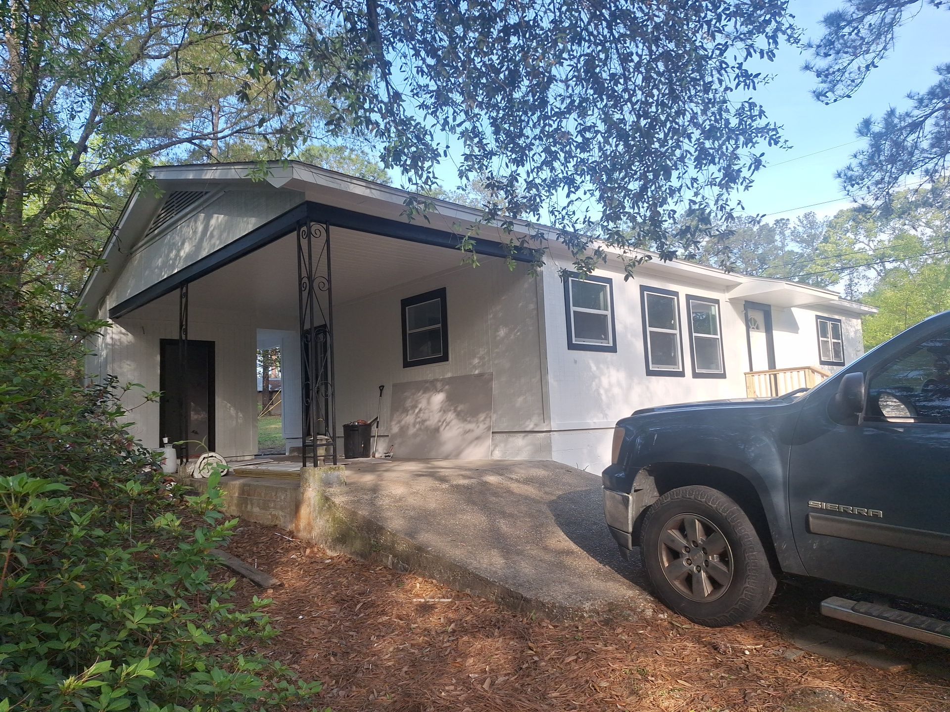Small house with front porch and parked dark pickup truck, surrounded by trees and shrubs