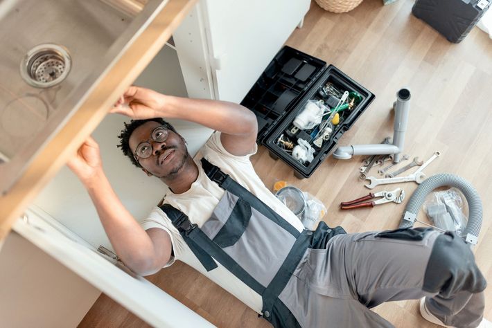 A Black plumber in overalls is fixing a sink, lying on the floor with a toolbox nearby.