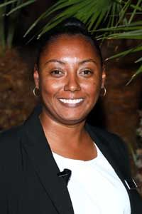 A woman in a black jacket and white shirt is smiling in front of a palm tree.