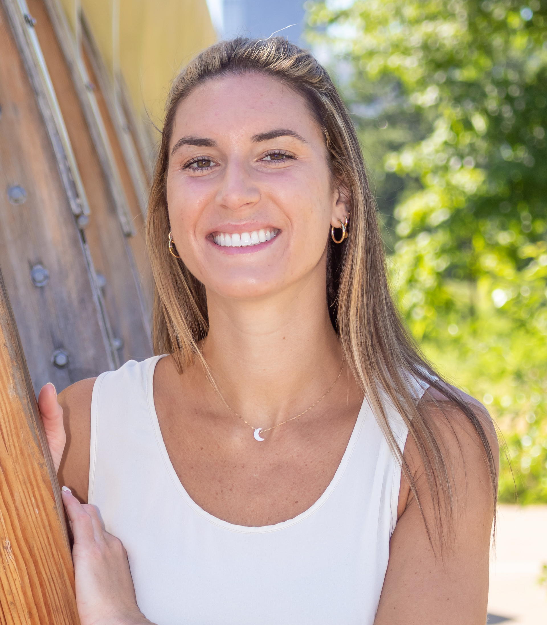 A woman in a white tank top is smiling while leaning against a wooden wall.