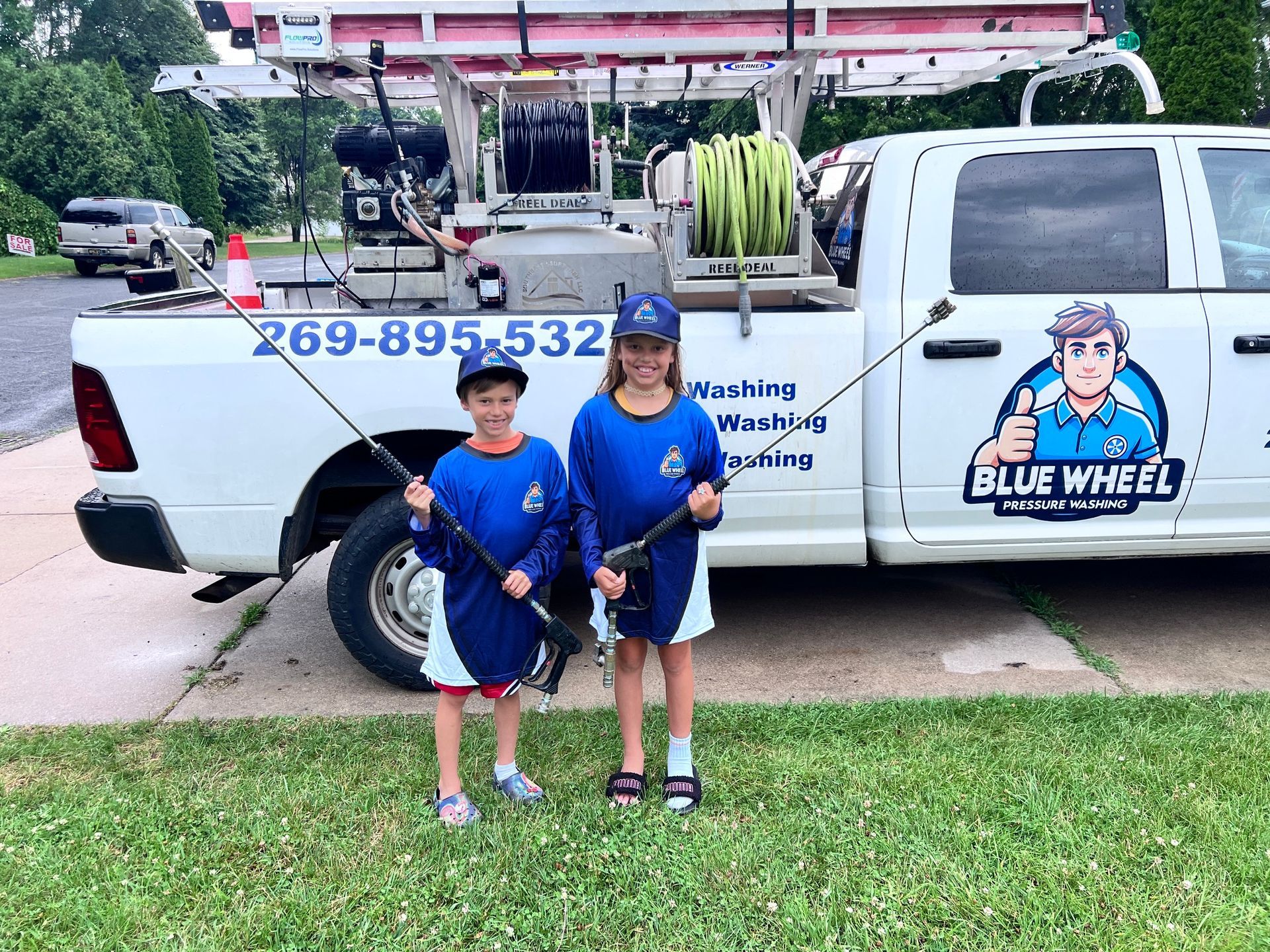 Two children in blue shirts standing in front of a white Blue Wheelz washing truck in a yard