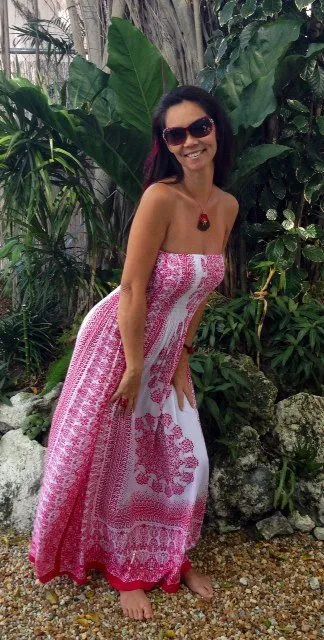 Woman in sunglasses wearing a pink patterned sundress, smiling and posing in a lush tropical garden.