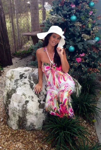 Woman in a pink floral dress and white hat sitting on a rock beside a decorated Christmas tree outdoors