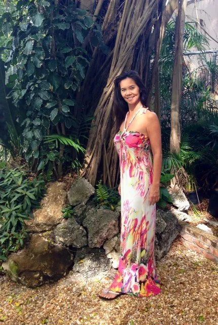 Woman in a pink floral dress standing by rocks and tropical greenery, smiling at the camera.