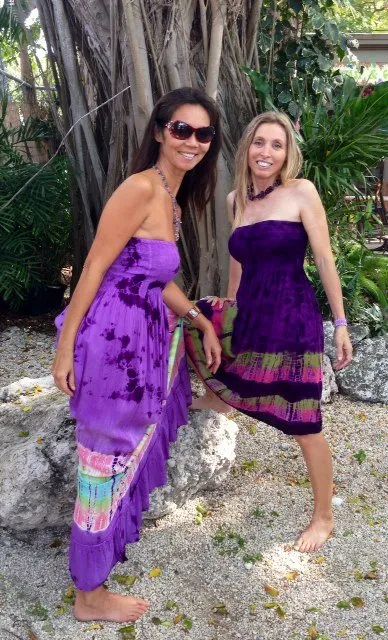 Two women in purple dresses pose barefoot on a rocky beach in front of tropical trees.