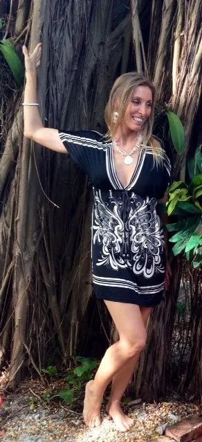 Woman in black patterned dress posing barefoot beside a large tree with tropical plants in the background