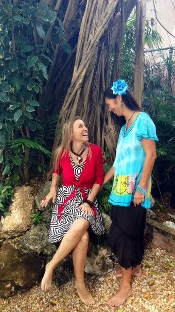 Two women in colorful dresses smiling beside a large tree in a tropical garden