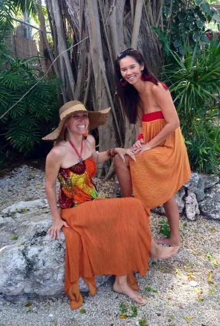 Two women in colorful dresses posing outdoors among tropical plants and rocks.