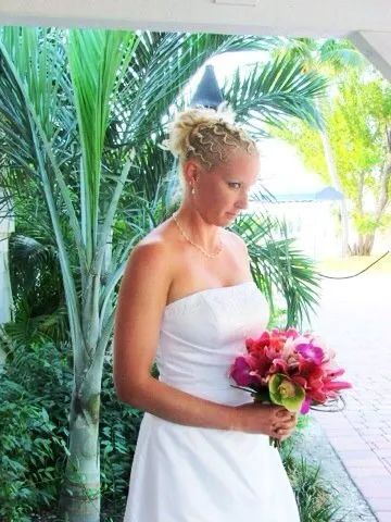 Woman in a white dress holding a pink bouquet, standing on a tropical walkway with palm trees.