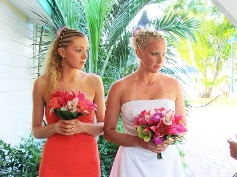 Two women holding bouquets, one in coral dress and one in white dress, standing in a tropical setting