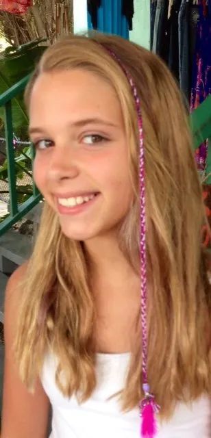 Smiling blonde child with pink-tipped braids, wearing a white top, in front of colorful decorations