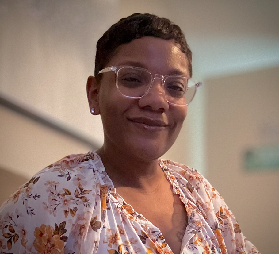 Smiling person wearing clear glasses and a floral blouse indoors.