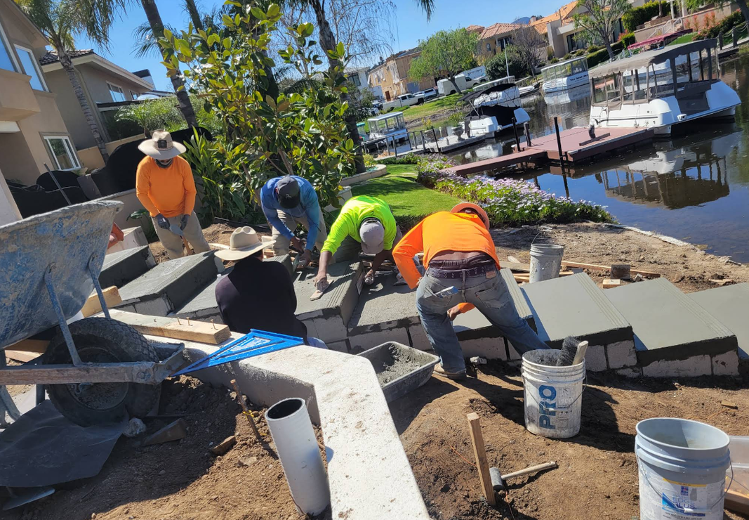 Workers pouring concrete along a waterfront walkway beside a canal and boats.