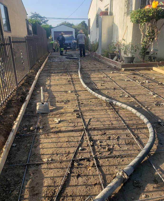 Construction work on a narrow outdoor path with workers, rebar, and a curved hose line beside buildings