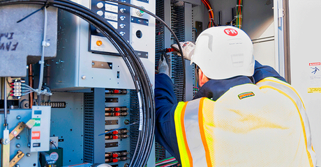 Technician in white hard hat inspecting electrical control panel with cables and wiring inside industrial cabinet