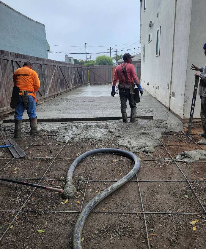 Two workers pouring concrete on a narrow outdoor path beside a building and fence