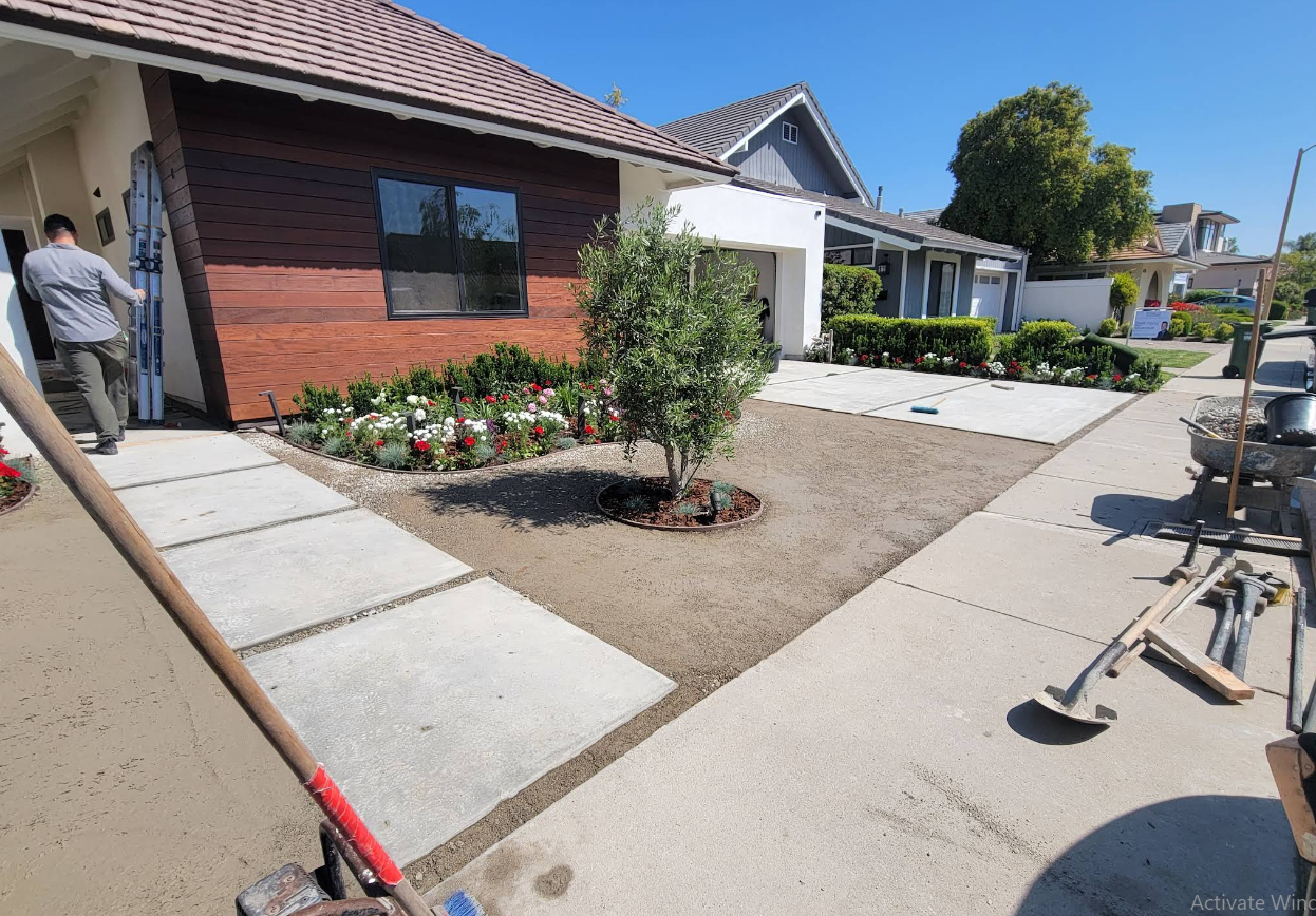 Residential front yard with a concrete walkway, flower beds, and a person on the left near a house under construction.