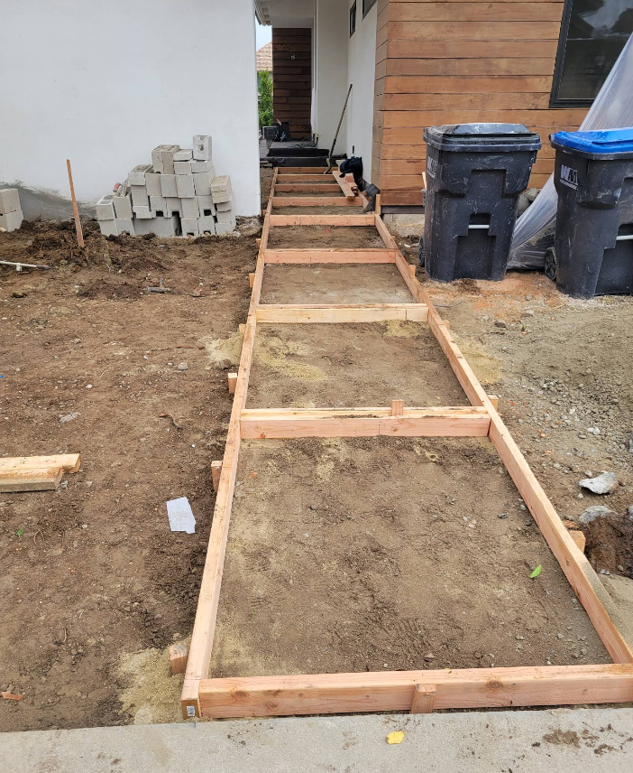 Wooden walkway frame under construction beside a house, with dirt fill and trash bins nearby
