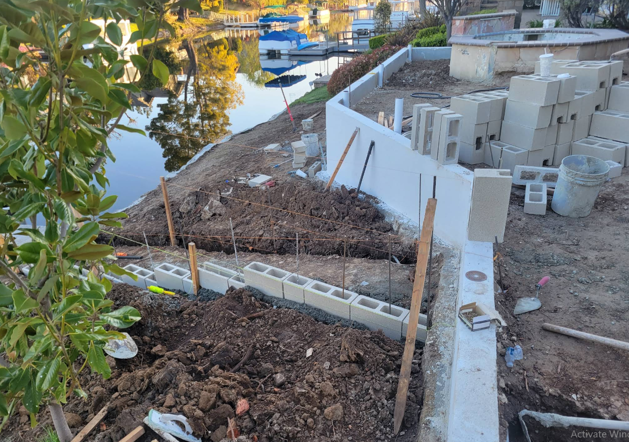 Garden beds beside a canal, with fresh soil, concrete blocks, and a partially built retaining wall.
